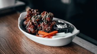 A white dish containing pieces of crispy, glazed chicken wings garnished with green herbs. Accompanied by carrot and celery sticks, along with a small black container of dipping sauce. The wooden table adds a rustic feel.