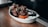 A white dish containing pieces of crispy, glazed chicken wings garnished with green herbs. Accompanied by carrot and celery sticks, along with a small black container of dipping sauce. The wooden table adds a rustic feel.