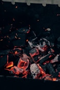 Lumps of charcoal are glowing with orange heat, surrounded by dark, charred pieces. Tiny sparks and embers can be seen floating upwards. The background is mainly dark, contrasting with the radiant glow of the burning coals.