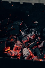 Lumps of charcoal are glowing with orange heat, surrounded by dark, charred pieces. Tiny sparks and embers can be seen floating upwards. The background is mainly dark, contrasting with the radiant glow of the burning coals.