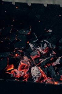 Lumps of charcoal are glowing with orange heat, surrounded by dark, charred pieces. Tiny sparks and embers can be seen floating upwards. The background is mainly dark, contrasting with the radiant glow of the burning coals.