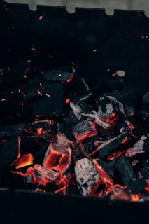 Lumps of charcoal are glowing with orange heat, surrounded by dark, charred pieces. Tiny sparks and embers can be seen floating upwards. The background is mainly dark, contrasting with the radiant glow of the burning coals.