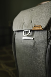 A close-up view of a well-crafted backpack made from dark gray fabric, featuring the brand name on a brown leather patch. A metal buckle near the top adds to its modern and functional design. The background is softly blurred, focusing attention on the backpack.