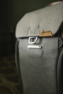 A close-up view of a well-crafted backpack made from dark gray fabric, featuring the brand name on a brown leather patch. A metal buckle near the top adds to its modern and functional design. The background is softly blurred, focusing attention on the backpack.