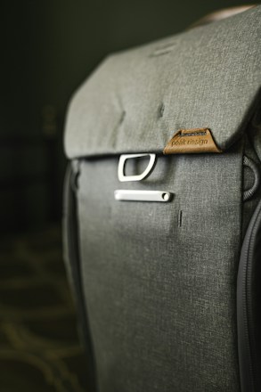 A close-up view of a well-crafted backpack made from dark gray fabric, featuring the brand name on a brown leather patch. A metal buckle near the top adds to its modern and functional design. The background is softly blurred, focusing attention on the backpack.