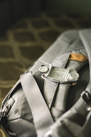 A close-up view of a grey backpack with a clip or mount attached to one of its straps. The material of the backpack appears durable and it features a small leather accent. The surroundings are softly lit, giving a sense of a comfortable setting.