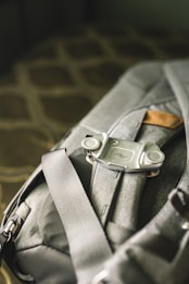 A close-up view of a grey backpack with a clip or mount attached to one of its straps. The material of the backpack appears durable and it features a small leather accent. The surroundings are softly lit, giving a sense of a comfortable setting.