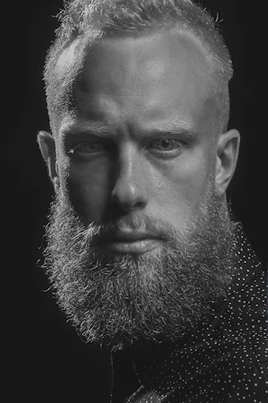 Portrait of a confident man with a sharp haircut and perfectly groomed beard against a deep black background with golden lighting.