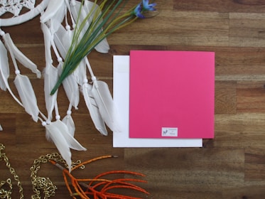 On a wooden surface, a bright pink card or envelope is positioned alongside a white envelope. To the left, there are decorative elements including white feathers on strings, green plant stems, and a purple flower. Orange twigs and a gold chain are also present, adding a dynamic contrast to the scene.