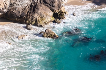 Turquoise waves gently crash against rugged, rocky cliffs along a serene coastline. Golden sand peeks through the edges where the water recedes, and white foam forms intricate patterns as the sea meets the shore. The sunlight casts a soft glow over the natural scenery, highlighting the textured surfaces of the rocks.