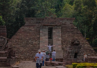 A stone pyramid structure with steps leading up, surrounded by lush green trees. A group of people dressed in traditional garments stand in front of the structure, with one person striking a pose with arms outstretched. The architecture suggests an ancient or historical site.