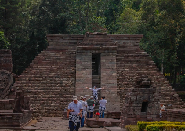 A stone pyramid structure with steps leading up, surrounded by lush green trees. A group of people dressed in traditional garments stand in front of the structure, with one person striking a pose with arms outstretched. The architecture suggests an ancient or historical site.