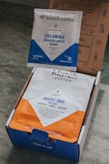 Two packages of coffee from Klatch Coffee are displayed. One is a small packet labeled 'Colombia Roasters United Blend' with a blue and white design. The other is a larger bag labeled 'Mocha Java Blend' with a blue and orange design. Both packages are situated inside an open cardboard box with printed text and graphics.
