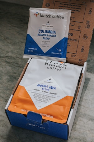 Two packages of coffee from Klatch Coffee are displayed. One is a small packet labeled 'Colombia Roasters United Blend' with a blue and white design. The other is a larger bag labeled 'Mocha Java Blend' with a blue and orange design. Both packages are situated inside an open cardboard box with printed text and graphics.