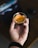 A hand holding a small glass filled with espresso, displaying a rich crema on top. The background shows a blurred image of a computer screen, suggesting a work or home office setting.