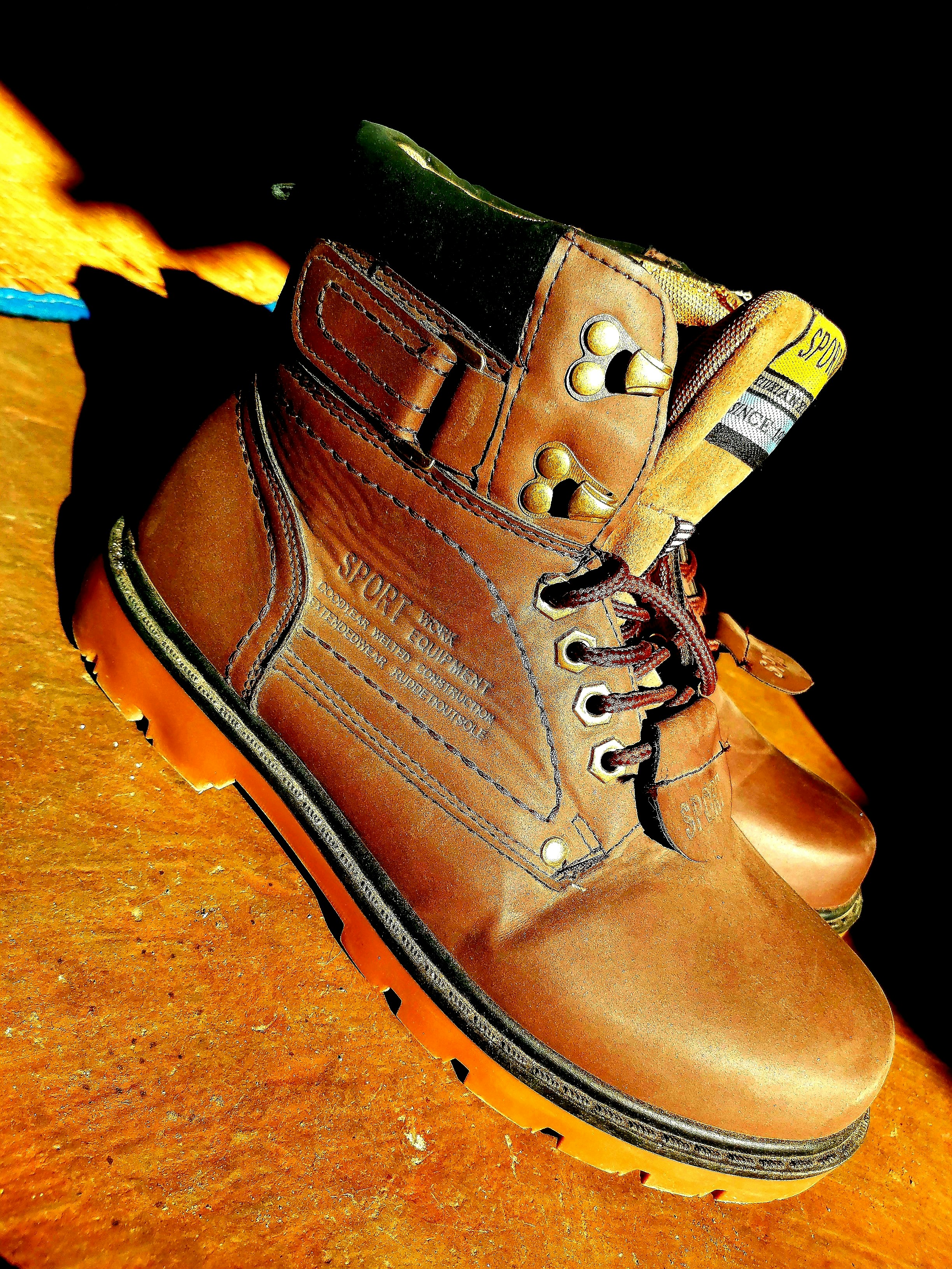 Rugged dark brown leather work boots with thick rubber soles on concrete industrial floor, dim workshop lighting