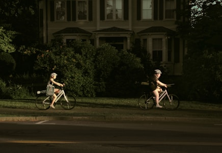 Two individuals are riding bicycles on a sidewalk beside a residential house surrounded by lush greenery. Both cyclists are wearing helmets and casual attire. The house in the background features multiple windows and is partially obscured by large bushes.