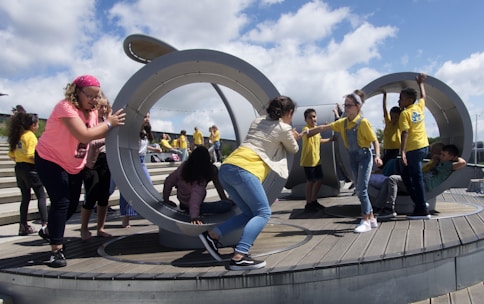 Children are actively playing in a large circular, metallic outdoor playground structure situated on a wooden deck. Some kids are pushing and interacting with the structure while others are climbing and sitting inside. A group of children in yellow shirts can be seen in the background on a stepped area.