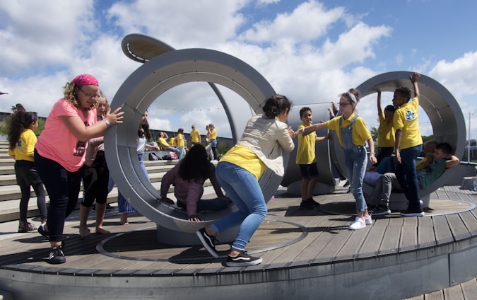 Children are actively playing in a large circular, metallic outdoor playground structure situated on a wooden deck. Some kids are pushing and interacting with the structure while others are climbing and sitting inside. A group of children in yellow shirts can be seen in the background on a stepped area.