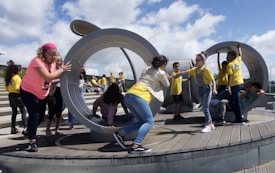 Children are actively playing in a large circular, metallic outdoor playground structure situated on a wooden deck. Some kids are pushing and interacting with the structure while others are climbing and sitting inside. A group of children in yellow shirts can be seen in the background on a stepped area.