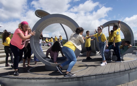 Children are actively playing in a large circular, metallic outdoor playground structure situated on a wooden deck. Some kids are pushing and interacting with the structure while others are climbing and sitting inside. A group of children in yellow shirts can be seen in the background on a stepped area.