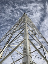 A tall metal tower structure extending into the sky, with workers climbing and working on it. The sky has light, wispy clouds providing a backdrop.