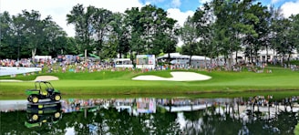 A lush green golf course scene with a sand bunker and a small pond in the foreground. In the distance, a large crowd of people can be seen gathered along the fairway, some seated on chairs. Behind them, scoreboards and tents are visible. A large John Deere vehicle is placed on a platform on the pond, its reflection mirrored in the water. Tall trees border the area, providing shade and a natural backdrop.