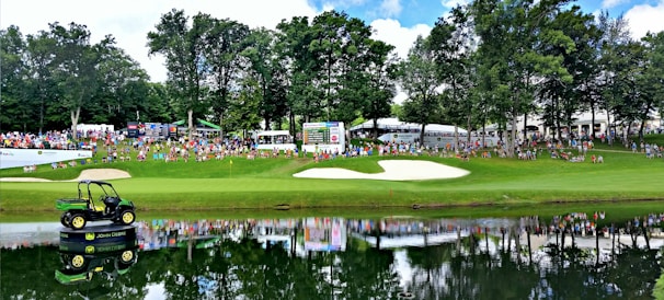 A lush green golf course scene with a sand bunker and a small pond in the foreground. In the distance, a large crowd of people can be seen gathered along the fairway, some seated on chairs. Behind them, scoreboards and tents are visible. A large John Deere vehicle is placed on a platform on the pond, its reflection mirrored in the water. Tall trees border the area, providing shade and a natural backdrop.