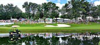 A lush green golf course scene with a sand bunker and a small pond in the foreground. In the distance, a large crowd of people can be seen gathered along the fairway, some seated on chairs. Behind them, scoreboards and tents are visible. A large John Deere vehicle is placed on a platform on the pond, its reflection mirrored in the water. Tall trees border the area, providing shade and a natural backdrop.