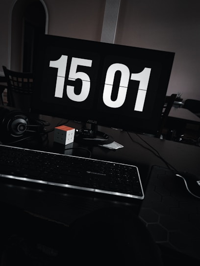 A dimly lit workspace featuring a computer monitor displaying the time 15:01 in large white digits. The setup includes a keyboard, a pair of headphones, a Rubik's cube, and a mouse pad on a dark desk.