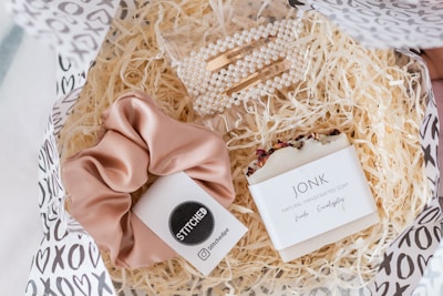 A gift box filled with shredded paper contains a few items. There is a pink satin scrunchie with a black and white label, a bar of natural handcrafted soap labeled Jonk with hints of red flower petals on top, and a set of pearl hair clips in a clear plastic bag.