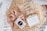 A gift box filled with shredded paper contains a few items. There is a pink satin scrunchie with a black and white label, a bar of natural handcrafted soap labeled Jonk with hints of red flower petals on top, and a set of pearl hair clips in a clear plastic bag.