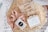 A gift box filled with shredded paper contains a few items. There is a pink satin scrunchie with a black and white label, a bar of natural handcrafted soap labeled Jonk with hints of red flower petals on top, and a set of pearl hair clips in a clear plastic bag.