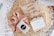 A gift box filled with shredded paper contains a few items. There is a pink satin scrunchie with a black and white label, a bar of natural handcrafted soap labeled Jonk with hints of red flower petals on top, and a set of pearl hair clips in a clear plastic bag.