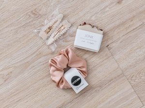 A set of pearl hair clips, a bar of natural handcrafted soap, and a beige satin scrunchie arranged on a light wood surface. The soap is labeled with 'JONK' and mentions scents of guido and eucalyptus.