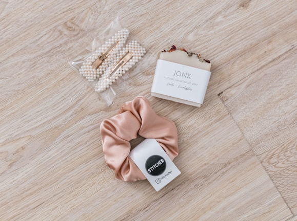 A set of pearl hair clips, a bar of natural handcrafted soap, and a beige satin scrunchie arranged on a light wood surface. The soap is labeled with 'JONK' and mentions scents of guido and eucalyptus.