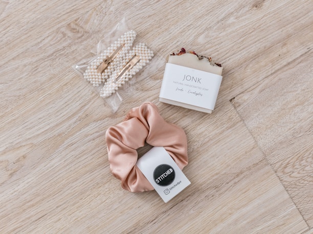 A set of pearl hair clips, a bar of natural handcrafted soap, and a beige satin scrunchie arranged on a light wood surface. The soap is labeled with 'JONK' and mentions scents of guido and eucalyptus.