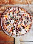 A delicious, freshly-baked pizza with a variety of toppings including green and red peppers, black olives, onions, pepperoni, ham, mushrooms, and melted cheese. The pizza is served on a wooden paddle placed on a rustic wooden table.