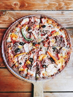 A delicious, freshly-baked pizza with a variety of toppings including green and red peppers, black olives, onions, pepperoni, ham, mushrooms, and melted cheese. The pizza is served on a wooden paddle placed on a rustic wooden table.