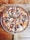 A delicious, freshly-baked pizza with a variety of toppings including green and red peppers, black olives, onions, pepperoni, ham, mushrooms, and melted cheese. The pizza is served on a wooden paddle placed on a rustic wooden table.