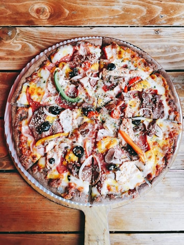 A delicious, freshly-baked pizza with a variety of toppings including green and red peppers, black olives, onions, pepperoni, ham, mushrooms, and melted cheese. The pizza is served on a wooden paddle placed on a rustic wooden table.