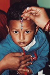 A young child in a blue hoodie is having decorative dots applied to their forehead with a cotton swab. The child's expression is serious and focused as someone carefully applies the colorful dots. Another hand gently supports the child.