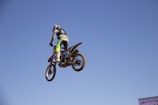 A rider in full gear launching a dirt bike off a dusty motocross jump under a bright blue sky.
