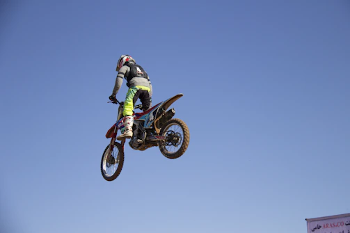 A motocross rider launching off a dirt jump, dust trailing behind under a clear blue sky.