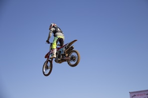 A rider in full gear launching a dirt bike off a dusty motocross jump under a bright blue sky.