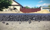 A close-up view of a freshly laid asphalt road, showcasing the rough texture and granular composition of the material. In the background, several structures are visible, including a beige house with a dark roof, a red fence, and construction mounds. The scene is set outdoors with clear weather and a blue sky featuring a few clouds.