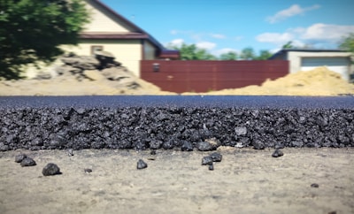 A close-up view of a freshly laid asphalt road, showcasing the rough texture and granular composition of the material. In the background, several structures are visible, including a beige house with a dark roof, a red fence, and construction mounds. The scene is set outdoors with clear weather and a blue sky featuring a few clouds.