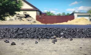 A close-up view of a freshly laid asphalt road, showcasing the rough texture and granular composition of the material. In the background, several structures are visible, including a beige house with a dark roof, a red fence, and construction mounds. The scene is set outdoors with clear weather and a blue sky featuring a few clouds.