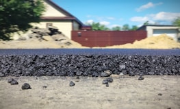 A close-up view of a freshly laid asphalt road, showcasing the rough texture and granular composition of the material. In the background, several structures are visible, including a beige house with a dark roof, a red fence, and construction mounds. The scene is set outdoors with clear weather and a blue sky featuring a few clouds.