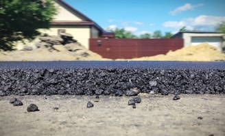 A close-up view of a freshly laid asphalt road, showcasing the rough texture and granular composition of the material. In the background, several structures are visible, including a beige house with a dark roof, a red fence, and construction mounds. The scene is set outdoors with clear weather and a blue sky featuring a few clouds.
